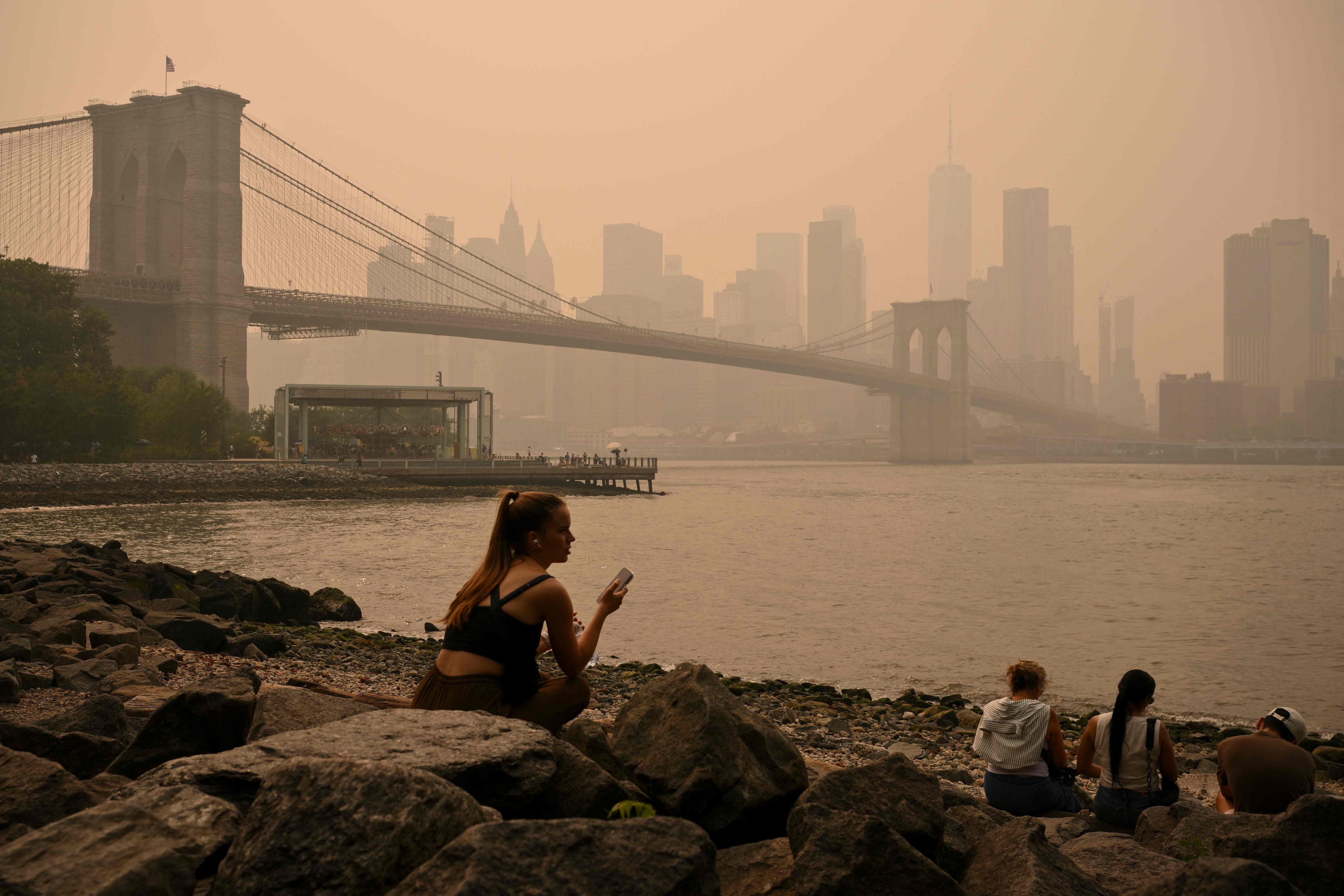 People sit near the Brooklyn Bridge as wildfire smoke from Canada causes a haze of smoke, July 16, 2026 in New York. Out-of-control wildfires raged Thursday in the Canadian province of Ontario, prompting evacuations and sending clouds of dangerous smoke toward the United States, where millions of people were exposed to unhealthy air.